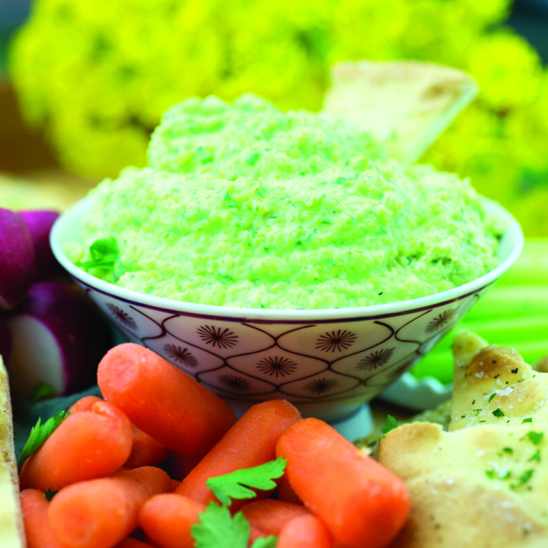 A bowl of creamy edamame hummus, garnished with olive oil, sesame seeds, and fresh herbs, served with pita bread and vegetable sticks.