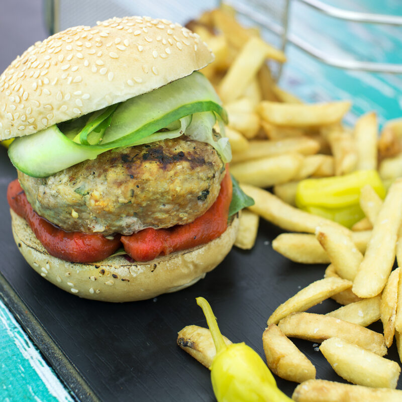 A Greek salad turkey burger served on a bun, topped with fresh vegetables, feta cheese, and olives.