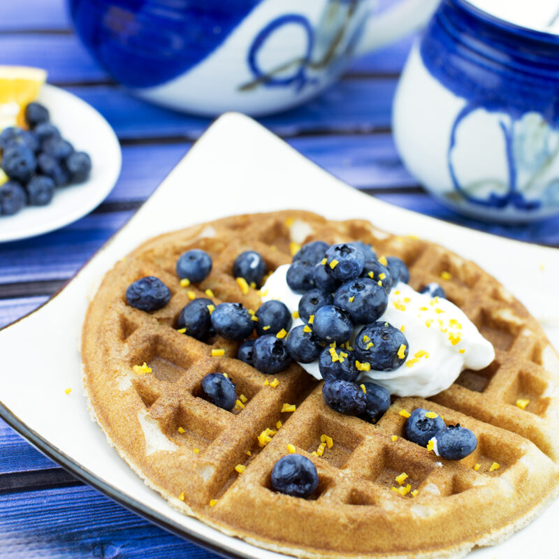 Sunshine blueberry waffles topped with fresh blueberries, lemon zest, and syrup on a plate.